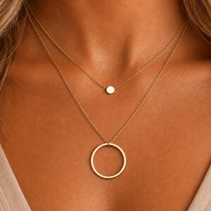 May include: A layered gold necklace featuring two delicate chains. The shorter chain holds a small, solid circular disc pendant, while the longer chain features a larger, open circular ring pendant. Both pendants are polished gold metal.