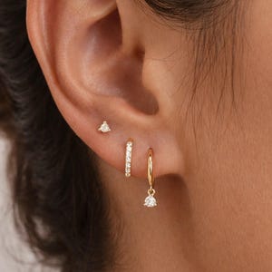 May include: A gold ear piercing set consisting of a small three-stone stud, a thin hoop earring with a row of clear pavé stones, and a plain gold hoop earring featuring a single dangling round clear stone charm, all worn in a single earlobe.