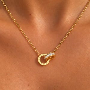 May include: A gold-toned chain necklace featuring two interlocking circular pendants. One pendant is a plain gold ring, while the other is a gold ring encrusted with a row of small, clear, sparkling stones along its outer edge.