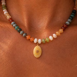 May include: A beaded necklace featuring a variety of small, smooth, multi-coloured stones in shades of green, orange, white, and dark grey. The necklace includes a central oval-shaped yellow pendant with a small, engraved sunburst design.