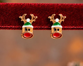 Puede incluir: Un par de pendientes de botón dorados con forma de cabeza de reno. Cada reno lleva un gorro verde con un pompón blanco, ojos negros y una nariz roja brillante. Los pendientes están sujetos a una tira horizontal de tela de terciopelo rojo.