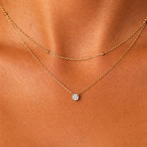 May include: A delicate, two-strand gold necklace. The top strand features small gold beads, while the lower strand holds a single, sparkling, round-cut diamond pendant. The necklace is displayed on a person's skin.
