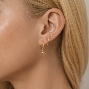 May include: Three gold-toned earrings consisting of one small star-shaped stud, one plain hoop, and one hoop featuring a dangling star charm. The jewelry pieces are polished and metallic, showcasing a consistent gold finish across all three items.