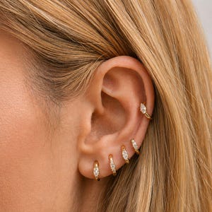 May include: A close-up of an ear adorned with multiple gold hoop earrings. Each earring features a small, oval-shaped, clear gemstone. The earrings vary in size and placement, creating a layered effect. The ear is set against a backdrop of blonde hair.