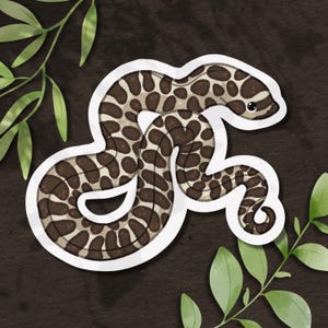 May include: A die-cut sticker featuring a cartoon snake with a light tan body and dark brown, irregular oval spots. The snake is coiled in a winding shape, has a small black eye, and is outlined with a thick white border against a dark background.