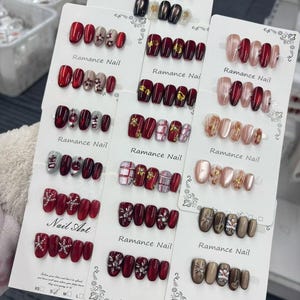 May include: A display of various press-on nail sets. The sets feature a range of colors, including red, nude, and black, with designs like glitter, floral accents, and rhinestones. Each set is displayed on a white card with the text "Ramance Nail".