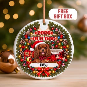 Puede incluir: Un adorno navideño redondo de cerámica blanca con una corona de pino verde, bayas rojas, piñas y luces. Un Cavalier King Charles Spaniel marrón con gorro de Papá Noel y bufanda de cuadros aparece con el texto 2026 THE YEAR WE WELCOMED OUR DOG y el nombre RUBY.