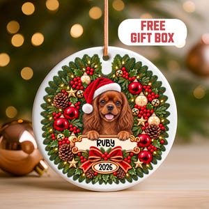 Op de afbeelding: Een ronde witte keramische kerstversiering met een bruine Cavalier King Charles-spaniël die een rode kerstmuts draagt. De hond is omringd door een krans van dennentakken, rode bessen, dennenappels en gouden sterren met de tekst RUBY en 2026.