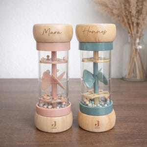 May include: Two wooden and clear plastic sensory toys. One is pink and the other is teal. Each has a wooden top with a name engraved on it. The base of each toy has the letter "J" engraved on it. The toys are filled with small colorful beads and a spinning mechanism.