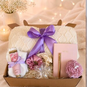 May include: A gift box containing a cream-colored knit blanket tied with a purple ribbon, a pink notebook labeled NOTES, a gold pen, a pink flower-shaped candle, two satin scrunchies, a clear flower-shaped accessory, and a tube of hand cream.