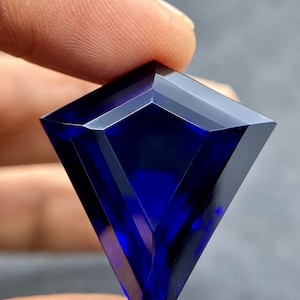 May include: A shield-shaped faceted gemstone with a deep, saturated royal blue colour. The stone features a geometric cut with sharp, clean edges and a pointed base, displaying high clarity and a smooth, reflective surface across its facets.