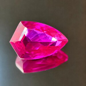 Puede incluir: Una piedra preciosa en forma de pera de color rosa intenso vibrante y saturado con una superficie facetada que crea reflejos de luz geométricos. La piedra es translúcida y presenta un corte angular nítido con un extremo puntiagudo y una base.