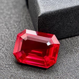 Könnte beinhalten: Ein leuchtend roter Edelstein im Smaragdschliff mit rechteckiger Form und facettierten Oberflächen, die das Licht reflektieren. Der Stein zeigt einen tiefen, gesättigten karmesinroten Farbton und liegt auf einer dunklen, strukturierten Oberfläche.