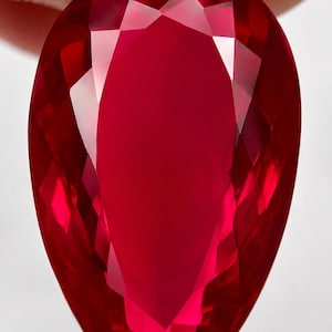 Puede incluir: Una gema de rubí facetada en forma de lágrima. La gema es de un color rojo intenso y saturado con múltiples facetas que reflejan la luz. La piedra se sostiene entre dos dedos.