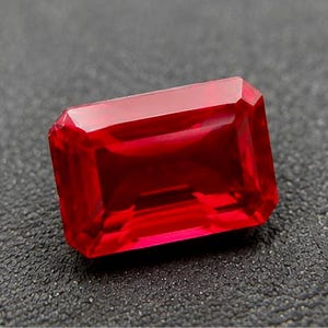 May include: A vibrant, deep red gemstone cut into a rectangular emerald shape. The stone features faceted edges that reflect light, showcasing a transparent, saturated crimson hue with a polished, smooth surface and sharp, defined geometric corners.