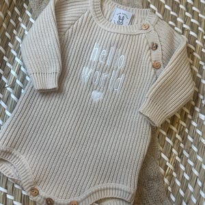 May include: A beige knitted baby romper with long sleeves and wooden buttons. The words "hello world" are embroidered in white on the front, along with a small heart. The romper has a round neckline and is made of a ribbed knit fabric.