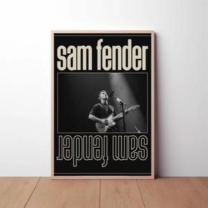 Puede incluir: P&oacute;ster en blanco y negro con el nombre "Sam Fender" en negrita y blanco en la parte superior e inferior. La imagen central es una fotograf&iacute;a en blanco y negro de un m&uacute;sico tocando la guitarra en el escenario bajo un foco.