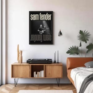 Puede incluir: P&oacute;ster enmarcado en blanco y negro con el texto "Sam Fender" arriba y abajo. Muestra a un m&uacute;sico tocando la guitarra. Cuelga sobre un mueble de madera con un tocadiscos.