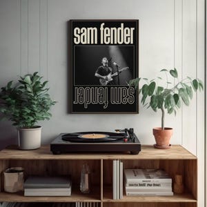 Puede incluir: Un p&oacute;ster en blanco y negro de Sam Fender cuelga sobre un tocadiscos en una estanter&iacute;a de madera. El p&oacute;ster presenta el nombre del artista y una imagen de &eacute;l tocando la guitarra. Hay plantas en macetas a ambos lados.