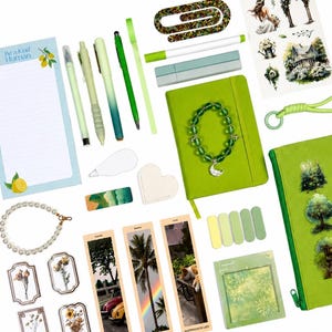 May include: A collection of stationery and accessories in shades of green, including a notepad with the text Be a Kind Human, pens, a green notebook, a beaded bracelet with a moon charm, bookmarks, stickers, a pearl bracelet, and a green pencil case.
