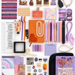 May include: A collection of stationery and craft supplies including an orange notebook, various pens, purple and orange washi tape, sticker sheets, bookmarks, a book tracker, a craft ink pad, a hand cream tube, a scrunchie, and decorative paper.
