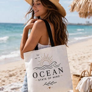 Puede incluir: Un bolso tote crema con correas negras, impreso con "OCEAN - STATE OF MIND" en negro y dorado, con un sol, olas y pájaros. El bolso se lleva en una playa.