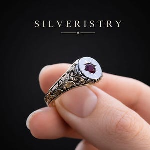 Puede incluir: Primer plano de un anillo de plata con una cara blanca y una gema roja oscura en forma de estrella. El anillo presenta intrincados diseños florales y de hojas. La palabra "SILVERISTRY" está en la parte superior.