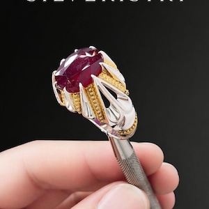 May include: A silver and gold ring with a large, oval-cut ruby gemstone. The ring has intricate detailing and is held by a silver tool. The word "SILVERISTRY" is displayed at the top of the image.