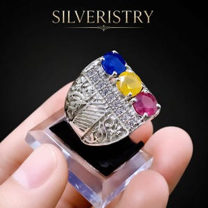 May include: A silver ring featuring the text SILVERISTRY at the top. The ring has a wide band with intricate scrollwork and a row of small clear stones. Three larger round faceted stones in blue, yellow, and red are set vertically on the face.