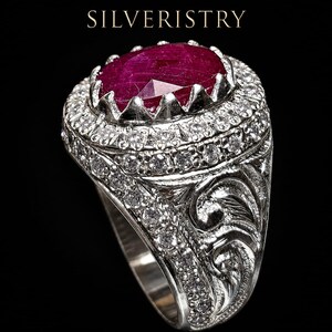 以下が含まれることがあります： 中央に大きな楕円形の深い赤色のルビーがセットされ、その周りを小さな透明な石が囲むシルバーリング。リングのバンド部分には複雑な彫刻と追加の透明な石が施されている。SILVERISTRYという文字が確認できる。