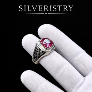 May include: A silver ring featuring a cushion-cut red gemstone set in a prong setting. The band displays intricate scrollwork engravings along the sides. The word SILVERISTRY is printed in white capital letters above the ring on a black background.