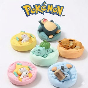 Op de afbeelding: Een verzameling Pokémon-beeldjes, elk rustend op een kleurrijk, pluche kussen. De beeldjes omvatten Snorlax, Eevee, Bulbasaur en anderen, met het Pokémon-logo bovenaan. De kussens zijn in verschillende pastelkleuren.