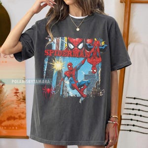Puede incluir: Camiseta gris oscuro con un gr&aacute;fico colorido de Spider-Man. El dise&ntilde;o incluye la palabra "SPIDERMAN" en rojo e ilustraciones de Spider-Man en acci&oacute;n contra un fondo de ciudad. Una prenda casual.