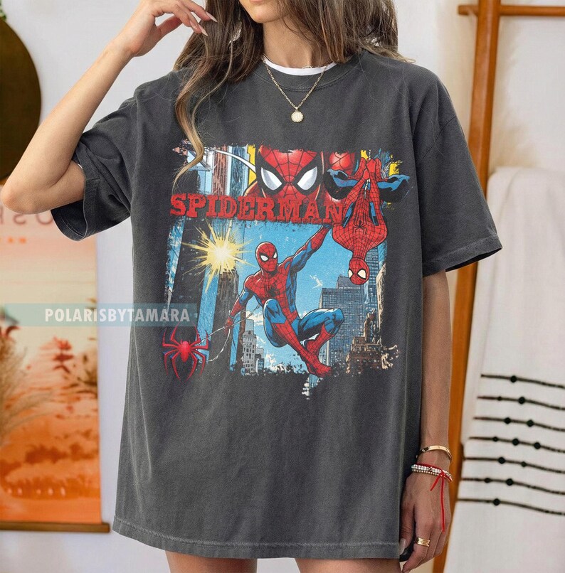 Puede incluir: Camiseta gris oscuro con un gr&aacute;fico colorido de Spider-Man. El dise&ntilde;o incluye la palabra "SPIDERMAN" en rojo e ilustraciones de Spider-Man en acci&oacute;n contra un fondo de ciudad. Una prenda casual.
