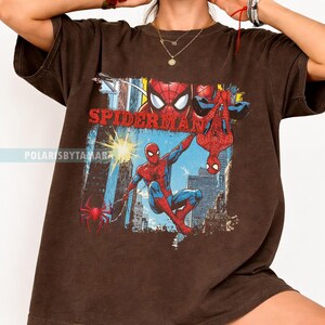 Puede incluir: Camiseta marr&oacute;n con un gr&aacute;fico colorido de Spider-Man en acci&oacute;n, con la palabra "SPIDERMAN" en letras rojas. El dise&ntilde;o incluye edificios de la ciudad y una telara&ntilde;a roja.
