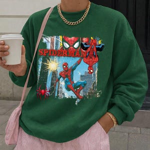 Puede incluir: Sudadera verde oscuro con un gr&aacute;fico colorido de Spider-Man. El dise&ntilde;o muestra a Spider-Man en acci&oacute;n con la palabra "SPIDERMAN" en rojo. El fondo presenta un paisaje urbano. La persona lleva una cadena y una pulsera doradas.