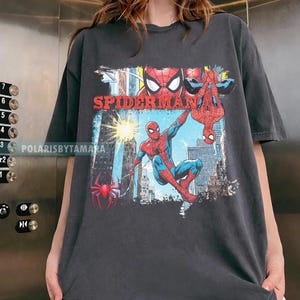 Puede incluir: Camiseta gris oscuro con un dise&ntilde;o de Spider-Man. El dise&ntilde;o incluye a Spider-Man en poses de acci&oacute;n, una ara&ntilde;a roja y la palabra "SPIDERMAN" en rojo. El fondo representa una ciudad.