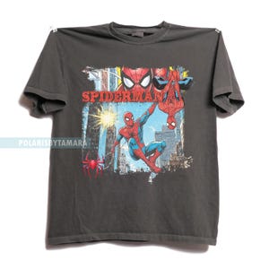 Puede incluir: Camiseta gris oscuro con un gr&aacute;fico colorido de Spider-Man en acci&oacute;n. El dise&ntilde;o incluye la palabra "SPIDERMAN" en rojo, junto con im&aacute;genes del superh&eacute;roe en varias poses contra un fondo de ciudad. Tambi&eacute;n es visible un logotipo de ara&ntilde;a roja.