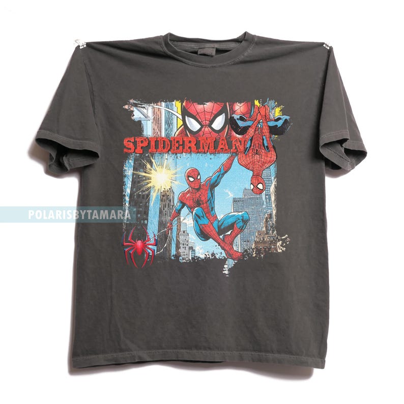 Puede incluir: Camiseta gris oscuro con un gr&aacute;fico colorido de Spider-Man en acci&oacute;n. El dise&ntilde;o incluye la palabra "SPIDERMAN" en rojo, junto con im&aacute;genes del superh&eacute;roe en varias poses contra un fondo de ciudad. Tambi&eacute;n es visible un logotipo de ara&ntilde;a roja.