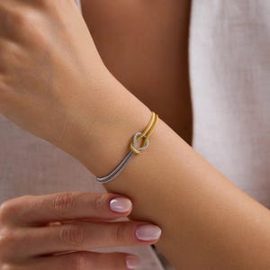 May include: A two-tone bracelet with a knot design. The bracelet has a silver-coloured chain and a gold-coloured knot. The bracelet is worn on a person's wrist.