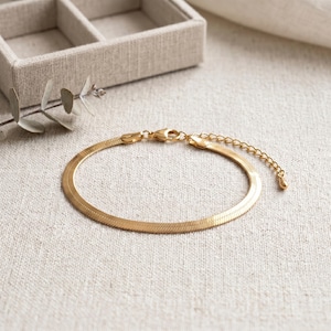 May include: A gold-colored herringbone bracelet with a lobster clasp and adjustable chain. The bracelet is displayed on a neutral-toned linen surface, with a small jewelry box in the background.