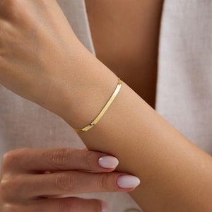 Puede incluir: Una pulsera dorada plana con diseño de espiga se lleva en la muñeca. La pulsera está hecha de pequeños eslabones rectangulares conectados. El tono de la piel es claro y las uñas están pintadas con un esmalte rosa claro.