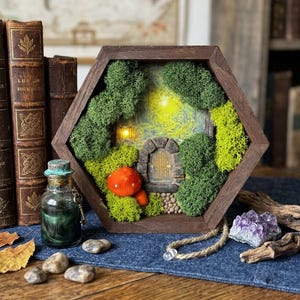 Puede incluir: Un marco hexagonal de madera alberga una escena de jardín de hadas en miniatura. La escena incluye una puerta de piedra, una seta roja y exuberante musgo verde. También hay una pequeña botella de vidrio con una llave y un cristal púrpura.