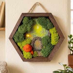 May include: A hexagonal wooden framed fairy garden scene. The scene features a miniature stone door, a red mushroom, and lush green moss. The background has a soft yellow glow, suggesting a light source within the miniature house.