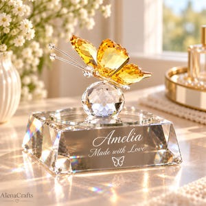 May include: A clear crystal butterfly with yellow wings perched on a crystal sphere atop a rectangular crystal base. The base is engraved with the name Amelia, the text Made with Love, and a small butterfly icon in a white script font.