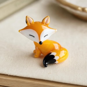 Könnte beinhalten: Eine kleine Glasfigur in Form eines Fuchses mit leuchtend orangefarbenem Körper sowie weißer Brust und weißem Gesicht. Der Fuchs hat spitze Ohren, eine schwarze Nase und geschlossene Augen. Der buschige Schwanz hat eine schwarz-weiße Spitze.