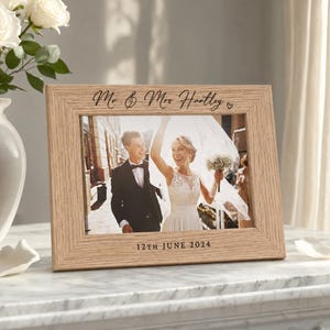 Puede incluir: Un marco de fotos rectangular de madera clara con el texto grabado Mr & Mrs Hartley en la parte superior y 12TH JUNE 2024 en la parte inferior. El marco contiene una fotografía de una pareja con ropa de boda, con la persona de vestido.