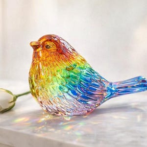 May include: A multi-coloured glass bird ornament, featuring shades of orange, yellow, green, and blue. A white rose lies beside the bird. The ornament is a decorative item, likely for display.