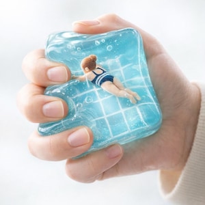 Könnte beinhalten: Eine Miniatur-Schwimmerin in einem blau-weiß gestreiften Badeanzug schwimmt in einer klaren, blauen, gelartigen Substanz. Das Gel hat ein Gittermuster, das einem Schwimmbecken ähnelt. Das Bild wird in einer Hand gehalten.