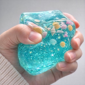 May include: A clear, square-shaped object filled with blue glitter and miniature seashells. The object is held in a hand, showcasing the translucent material and the decorative elements within. The seashells are in various colors.